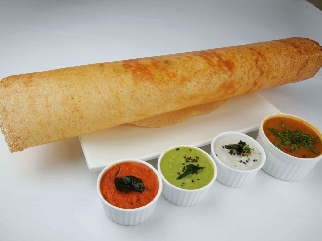 a dosa on a plate with sambar and chutneys in serving bowls