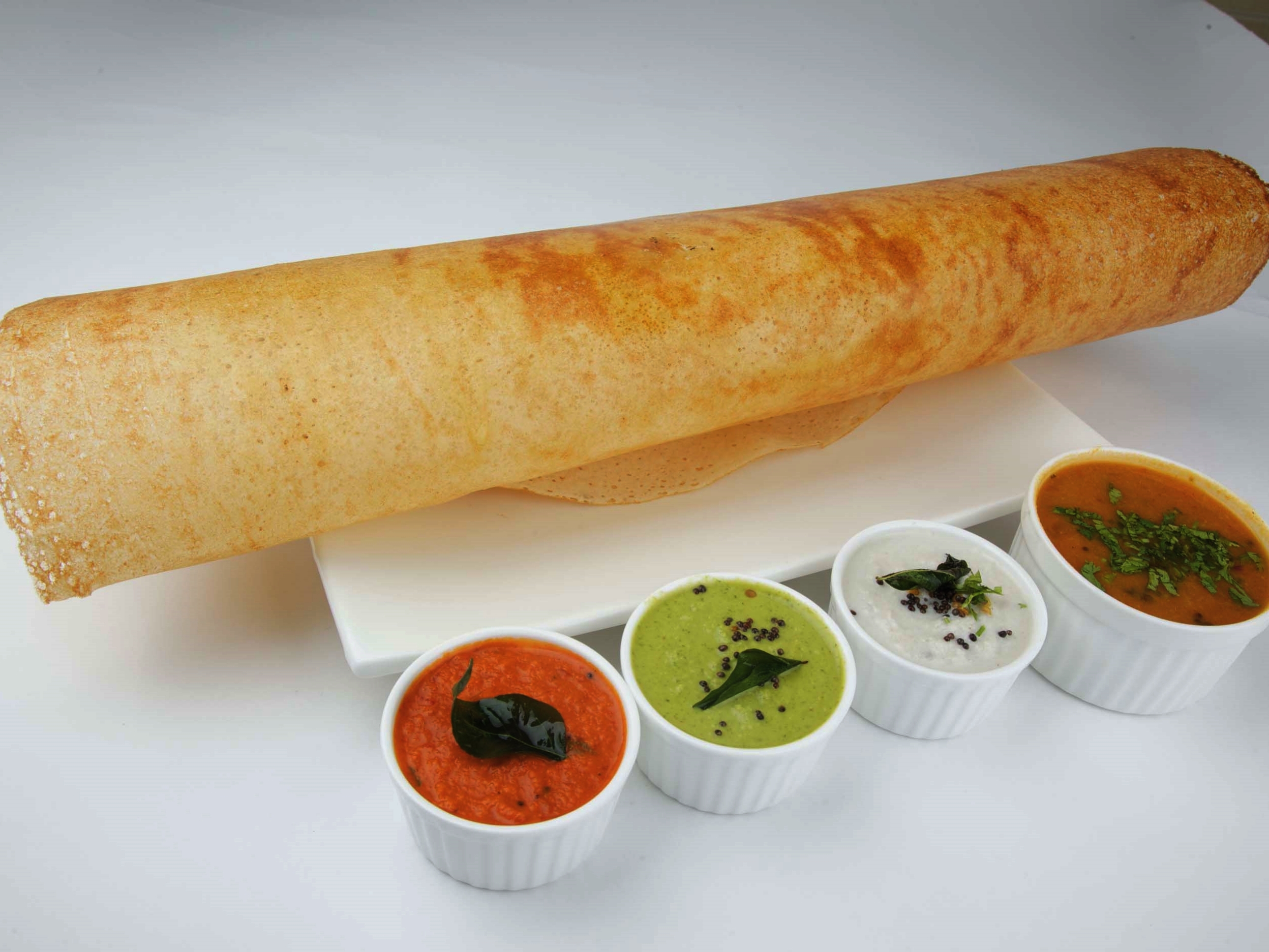 a dosa on a plate with sambar and chutneys in serving bowls