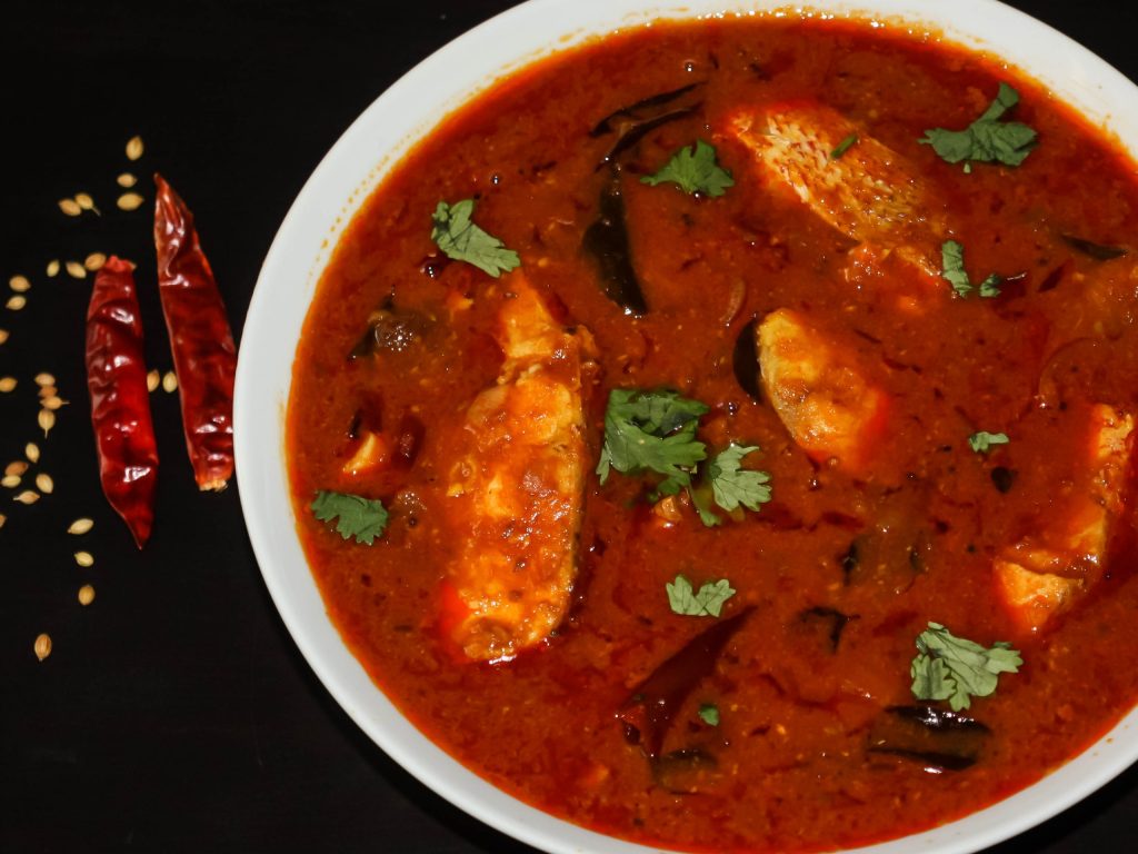 A serving bowl of chennai fish curry, with some dried red chillies on the side.